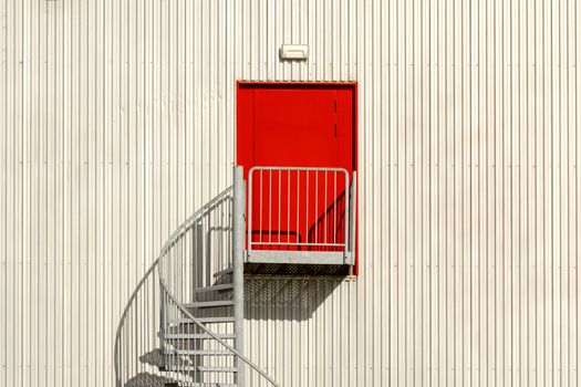 Minimalist architecture with a spiral staircase and vibrant red door against a corrugated metal wall.